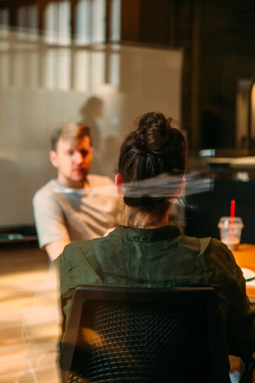 Two colleagues in conversation across a table in warm light, one seen from behind.